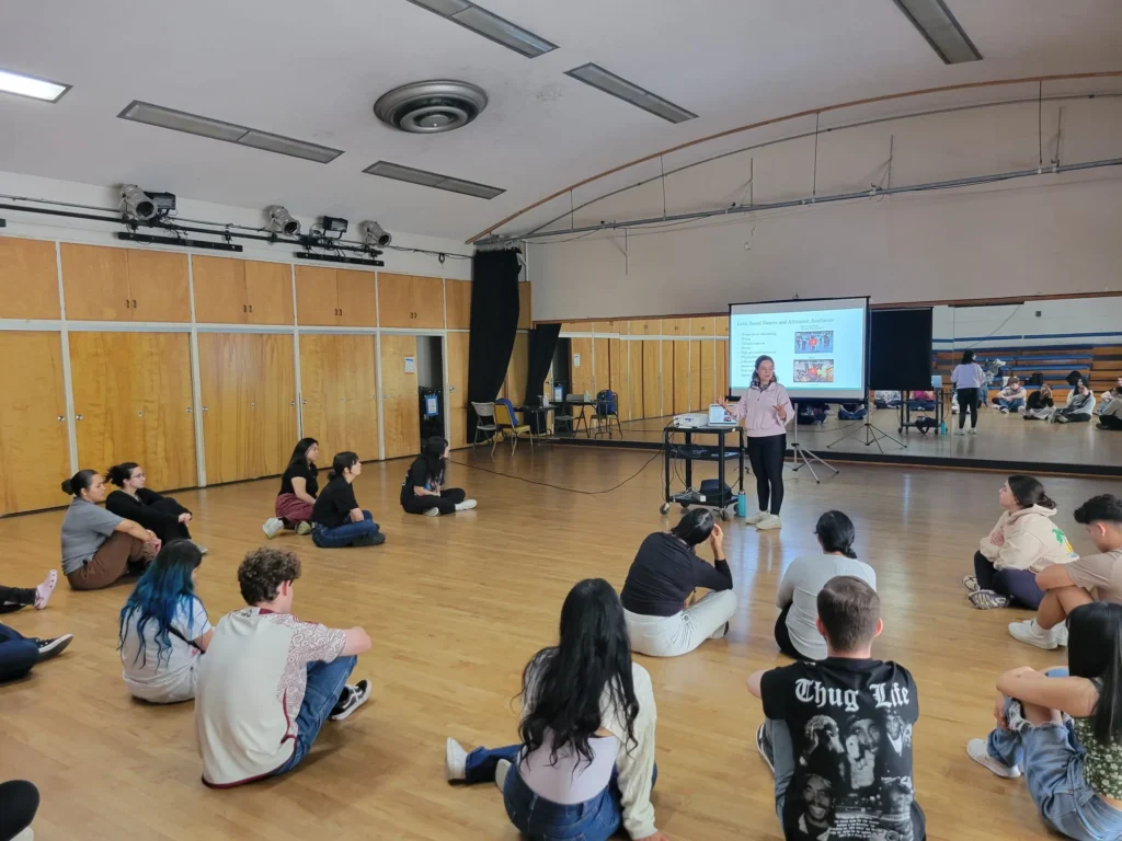 Cinthia Durán presenting to a seated group in a dance studio, mirrored wall behind